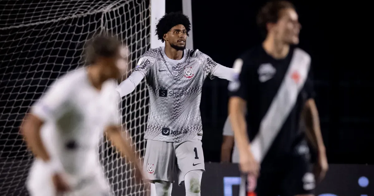 Jogador do Corinthians, de uniforme cinza com o número 1, gesticula em campo ao lado de uma rede de gol, enquanto atletas de camisa preta e branca aparecem desfocados em primeiro plano