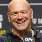 Homem careca e sorridente segura microfone com logo do UFC, diante de painel com patrocinadores ao fundo