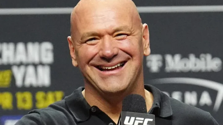 Homem careca e sorridente segura microfone com logo do UFC, diante de painel com patrocinadores ao fundo
