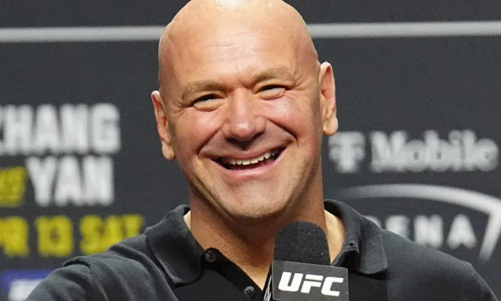 Homem careca e sorridente segura microfone com logo do UFC, diante de painel com patrocinadores ao fundo