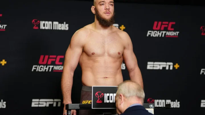 Lutador sem camisa, de barba, sobe em balança oficial durante pesagem do UFC Fight Night, com painéis de patrocinadores ao fundo, enquanto um oficial observa o peso
