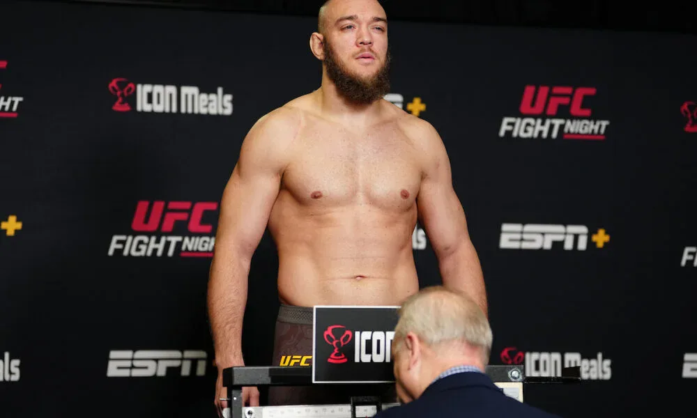 Lutador sem camisa, de barba, sobe em balança oficial durante pesagem do UFC Fight Night, com painéis de patrocinadores ao fundo, enquanto um oficial observa o peso