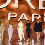 Grupo de mulheres desfila de mãos dadas em passarela com fundo laranja iluminado durante apresentação de moda em Paris