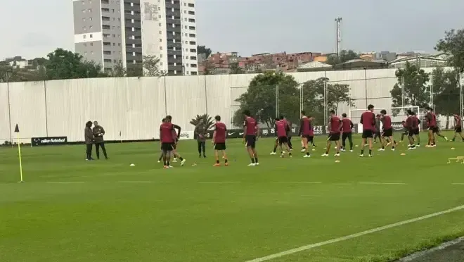Jogadores de camisa vermelha e short preto treinam em campo gramado, com prédios ao fundo, sob o olhar de membros da comissão técnica