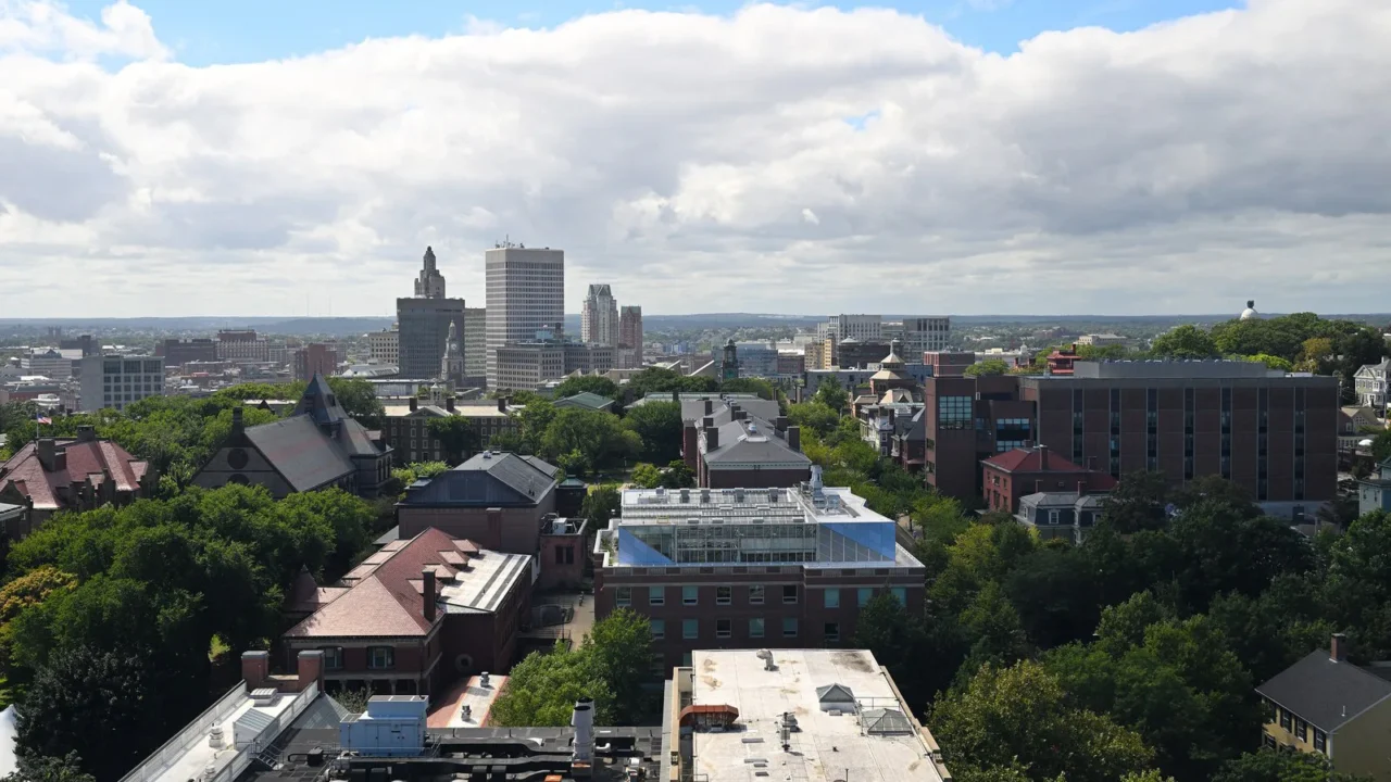 Foto de arquivo de Providence, em Rhode Island, vista do alto, com árvores, prédios baixos de tijolo e o centro da cidade ao fundo, sob céu azul com nuvens