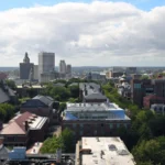 Foto de arquivo de Providence, em Rhode Island, vista do alto, com árvores, prédios baixos de tijolo e o centro da cidade ao fundo, sob céu azul com nuvens