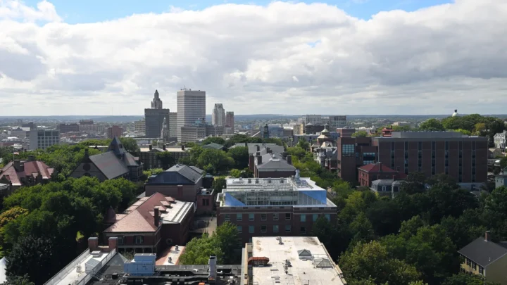 Foto de arquivo de Providence, em Rhode Island, vista do alto, com árvores, prédios baixos de tijolo e o centro da cidade ao fundo, sob céu azul com nuvens