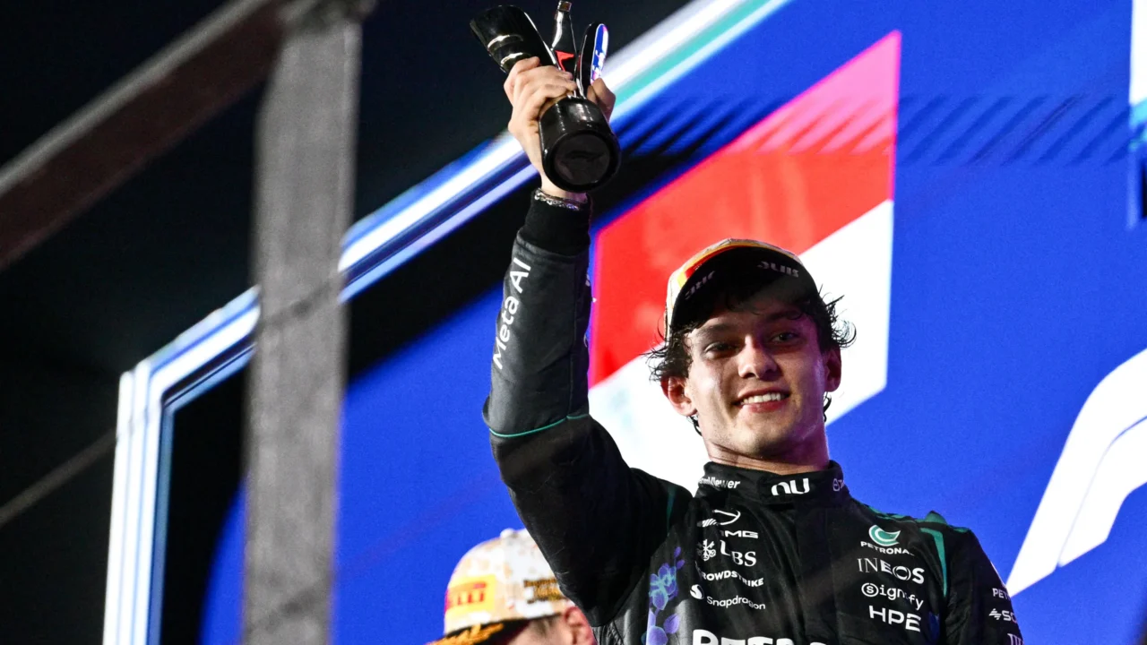 Piloto de macacão preto da Mercedes ergue troféu no pódio, sorridente, com patrocinadores como Petronas e INEOS ao fundo