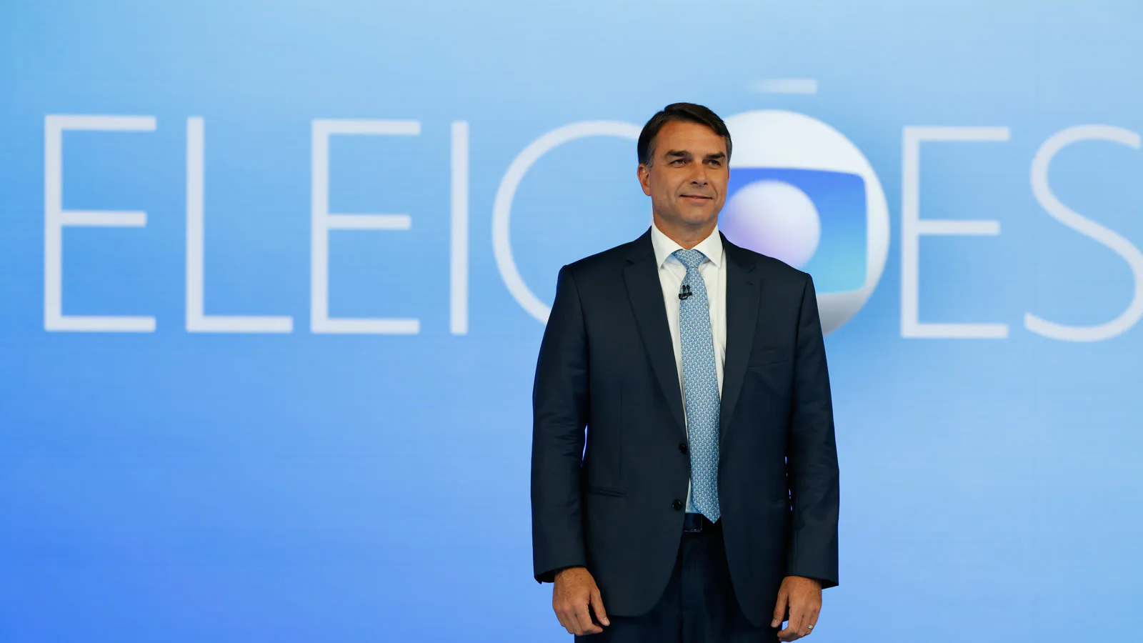 Flávio Bolsonaro, de terno azul-escuro e gravata azul-clara, de pé diante do painel "Eleições" no estúdio do debate da TV Globo, no Rio de Janeiro, em 23 de outubro de 2026