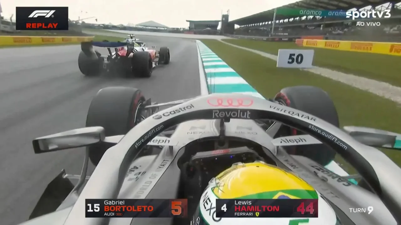 Piloto em carro branco com capacete amarelo e verde segue em pista molhada durante treino na Malásia, com outro veículo atrás em curva do circuito