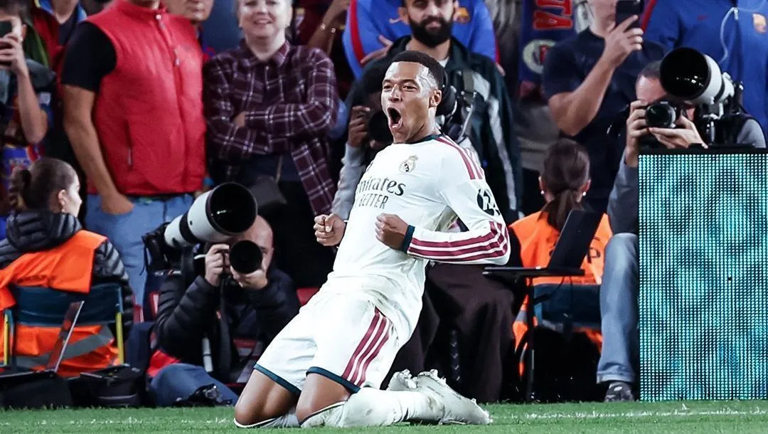 Jogador de camisa branca do Real Madrid ajoelha-se no gramado e comemora com os braços cerrados, cercado por fotógrafos na beira do campo