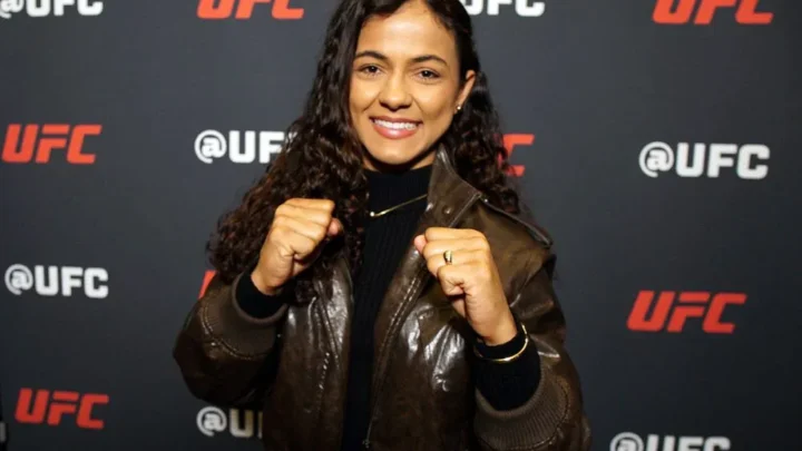 Mulher sorridente de jaqueta de couro marrom, com os punhos cerrados em pose de luta, diante de painel com a marca UFC