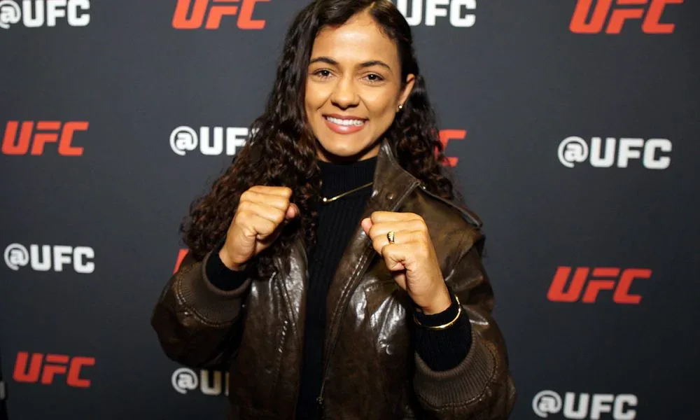 Mulher em evento do UFC posa com os punhos cerrados diante de painel da organização, usando jaqueta de couro preta