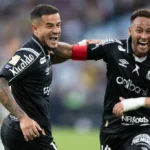 Dois jogadores do Santos com camisas pretas comemoram com muita empolgação, um deles com braçadeira vermelha, em campo de futebol