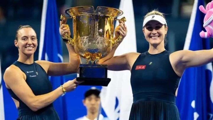 Duas tenistas sorridentes erguem um grande troféu dourado em uma premiação, com bandeiras azuis e brancas ao fundo