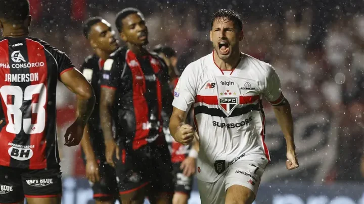 Jogador do São Paulo, de camisa branca, comemora com os punhos cerrados enquanto corre sob a chuva, com jogadores do Vitória de uniforme preto e vermelho ao fundo