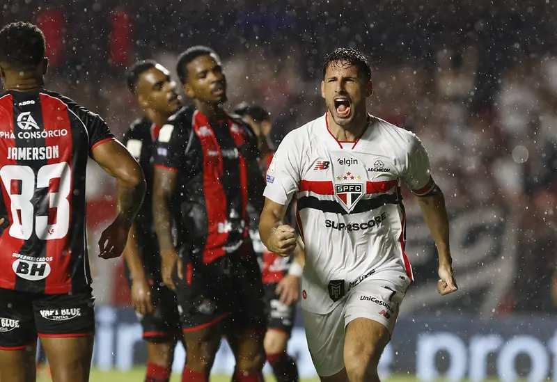 Jogador do São Paulo, de camisa branca, comemora com os punhos cerrados enquanto corre sob a chuva, com jogadores do Vitória de uniforme preto e vermelho ao fundo