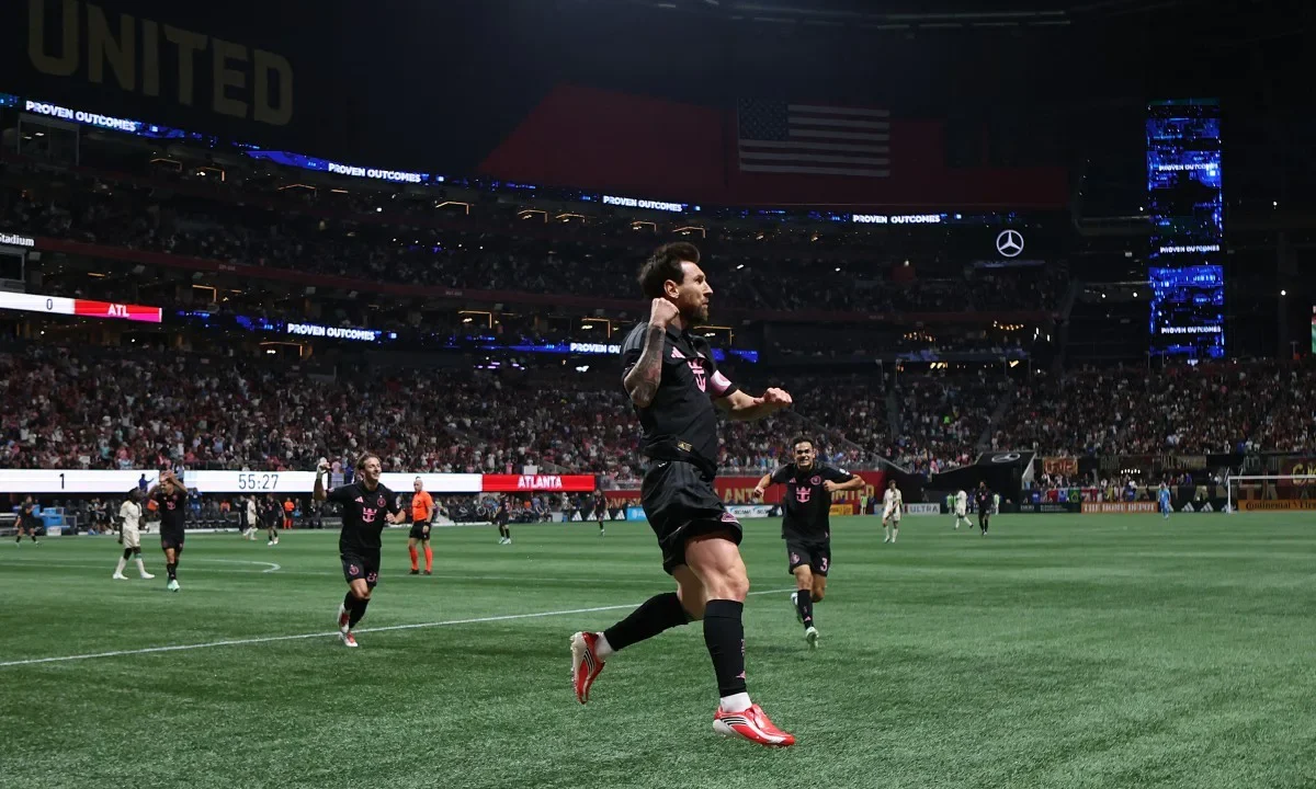 Jogador de camisa preta e vermelha corre comemorando com o braço erguido em estádio lotado, com placar mostrando 1 a 0 para o Atlanta e 55:27 no relógio