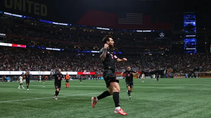 Jogador de camisa preta e vermelha corre comemorando com o braço erguido em estádio lotado, com placar mostrando 1 a 0 para o Atlanta e 55:27 no relógio