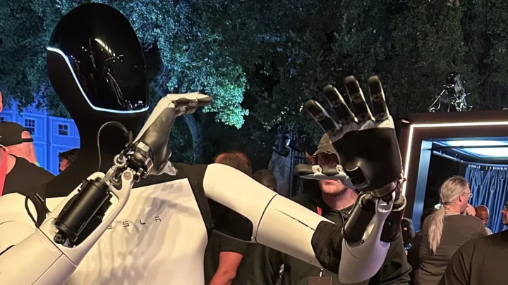 Robô humanoide Optimus, da Tesla, com capacete preto e corpo branco, acena com as mãos no meio do público num evento da empresa nos estúdios da Warner Bros., na Califórnia, em outubro de 2024 (imagem de arquivo)