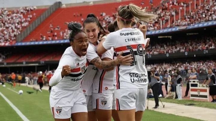 Jogadoras do São Paulo em uniforme branco celebram abraçadas em campo durante partida em estádio repleto de torcedores
