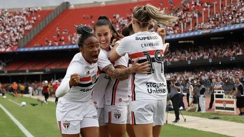 Jogadoras do São Paulo em uniforme branco celebram abraçadas em campo durante partida em estádio repleto de torcedores