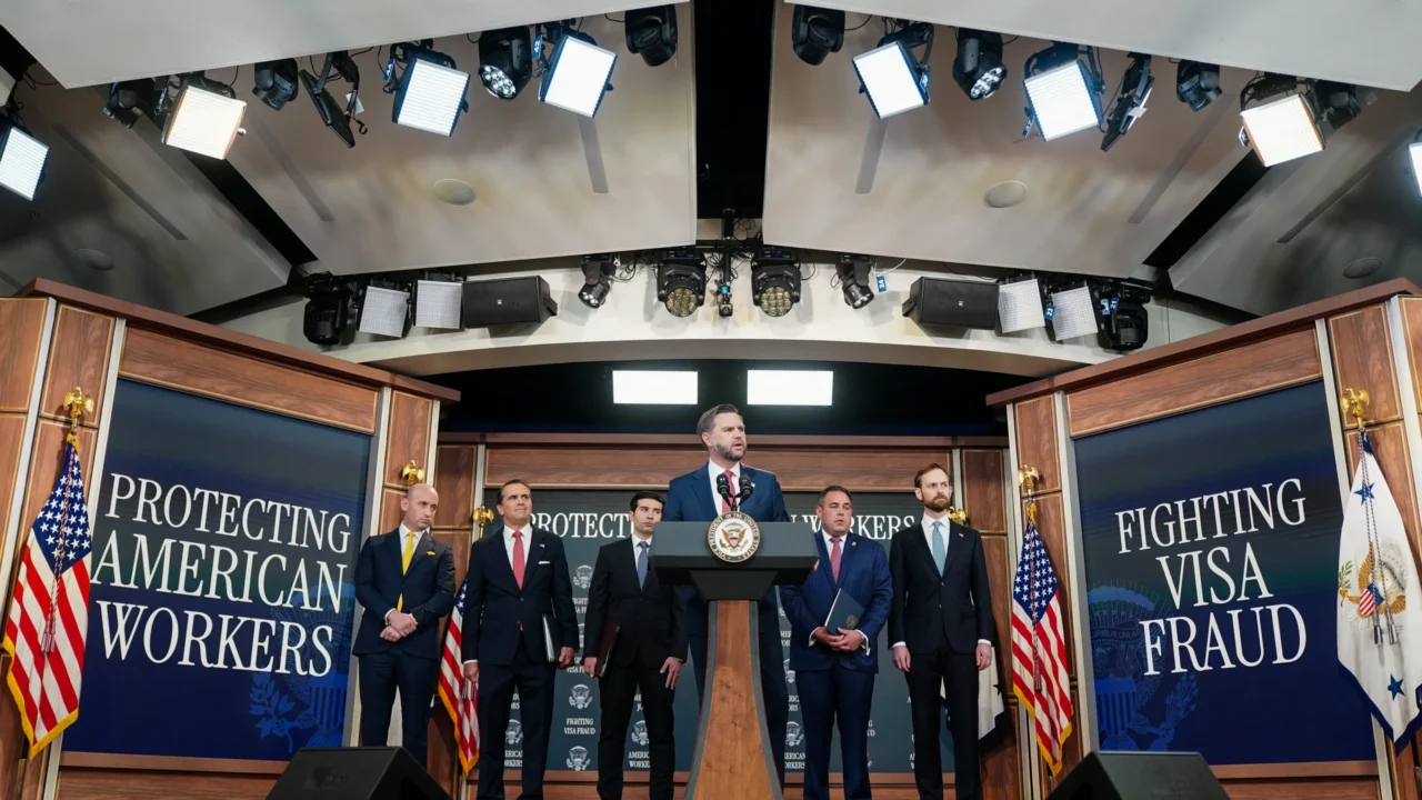 Vice-presidente dos EUA, JD Vance, discursa em pódio com a insígnia oficial, ladeado por cinco homens de terno em cenário com as frases "Protecting American Workers" e "Fighting Visa Fraud