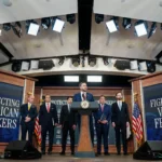 Vice-presidente dos EUA, JD Vance, discursa em pódio com a insígnia oficial, ladeado por cinco homens de terno em cenário com as frases "Protecting American Workers" e "Fighting Visa Fraud