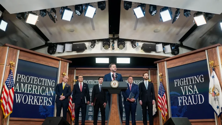 Vice-presidente dos EUA, JD Vance, discursa em pódio com a insígnia oficial, ladeado por cinco homens de terno em cenário com as frases "Protecting American Workers" e "Fighting Visa Fraud