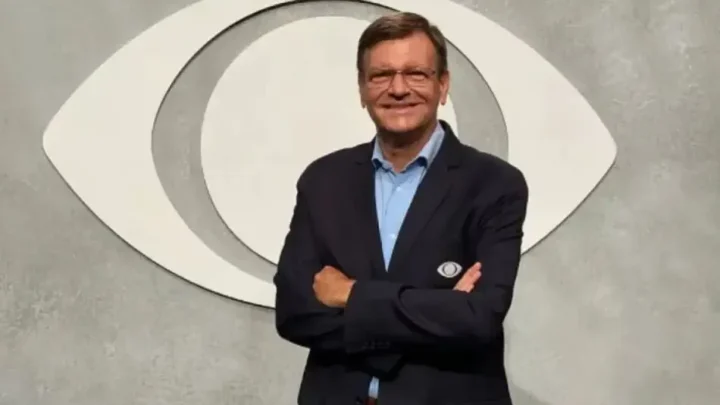Homem de terno e óculos com os braços cruzados posa diante de um logotipo de emissora de televisão