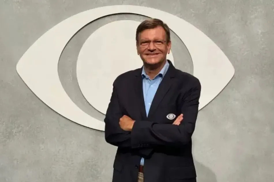 Homem de terno e óculos com os braços cruzados posa diante de um logotipo de emissora de televisão