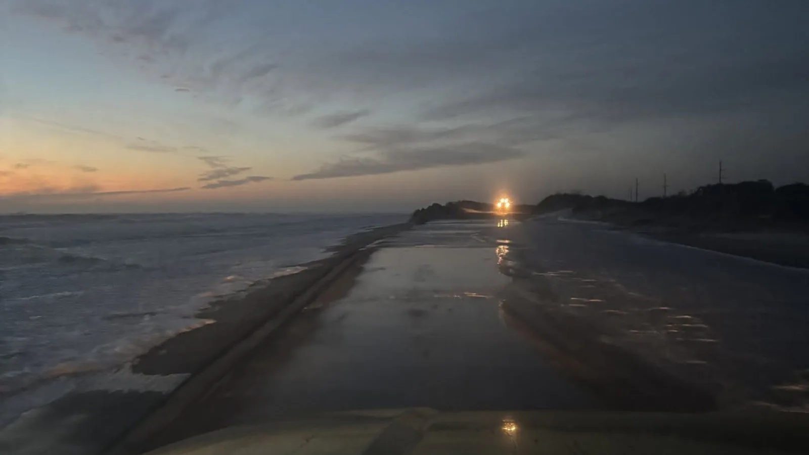 Trecho da NC 12 coberto de água e areia ao entardecer, visto de dentro de um veículo de trabalho