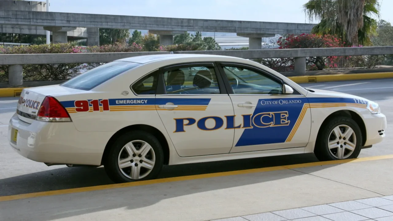 Viatura branca da polícia da cidade de Orlando, com faixas azuis e a palavra "Police" na porta, estacionada junto a um viaduto