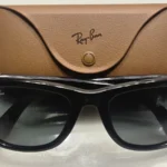 Óculos inteligentes Ray-Ban Meta de primeira geração, de armação preta, apoiados diante do estojo de couro marrom com a marca Ray-Ban (imagem ilustrativa)