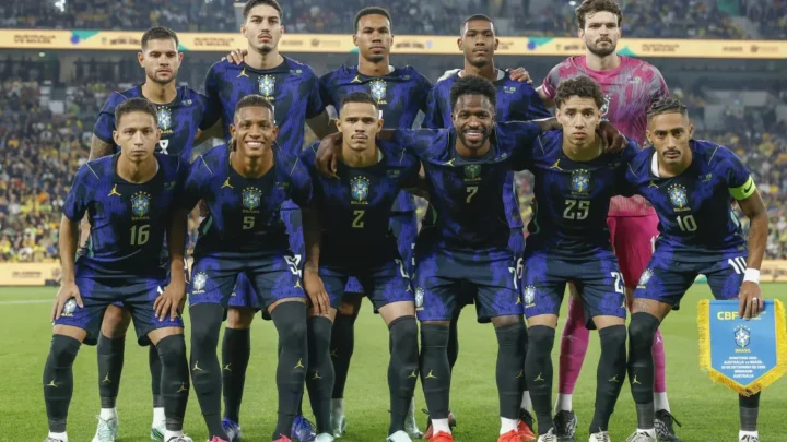 Seleção brasileira de futebol posicionada no campo antes de partida, com jogadores em uniforme azul e um goleiro em uniforme rosa ao fundo