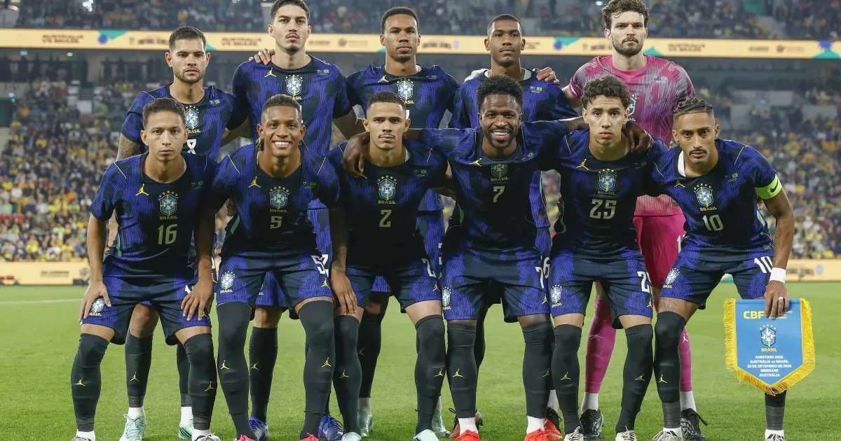 Seleção brasileira de futebol posicionada no campo antes de partida, com jogadores em uniforme azul e um goleiro em uniforme rosa ao fundo