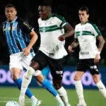 Jogadores de camisas azul e branca e verde e branca disputam a bola em campo, com placas de publicidade ao fundo