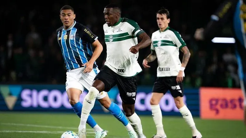 Jogadores de camisas azul e branca e verde e branca disputam a bola em campo, com placas de publicidade ao fundo