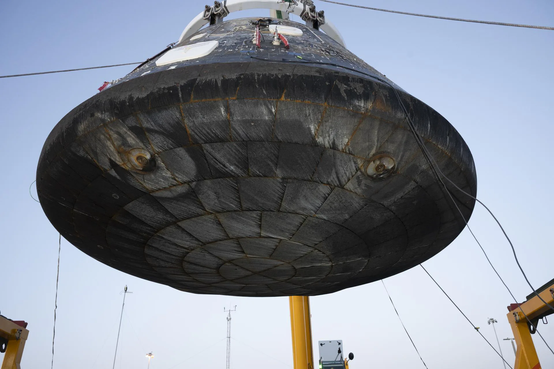 O módulo de tripulação Orion da Artemis II é içado de seu dispositivo de recuperação para inspeção e registro de imagens de engenharia, após ser descarregado do USS John P. Murtha na Base Naval de San Diego, em 18 de abril de 2026. As áreas recuadas, ou círculos concêntricos menores, vistas no escudo térmico, estão associadas aos parafusos de separação que conectam o módulo de tripulação ao módulo de serviço. Os padrões circulares maiores se formam devido ao material do bloco de compressão usado nessa localização da espaçonave, criando perturbações no fluxo causadas pelo fluxo aerodinâmico e térmico extremo durante a reentrada. Isso provoca aumento localizado do aquecimento e, por isso, a espessura do material ablativo Avcoat é aumentada nessas áreas por projeto.