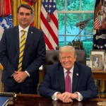 Um homem de terno azul-marinho e gravata listrada em amarelo e verde fica de pé ao lado de outro homem sentado atrás de uma mesa de madeira, em um escritório com bandeiras dos Estados Unidos e estátua de bronze ao fundo