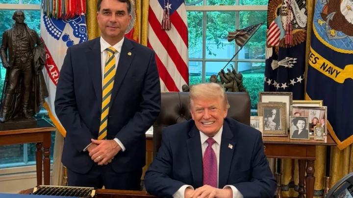 Um homem de terno azul-marinho e gravata listrada em amarelo e verde fica de pé ao lado de outro homem sentado atrás de uma mesa de madeira, em um escritório com bandeiras dos Estados Unidos e estátua de bronze ao fundo