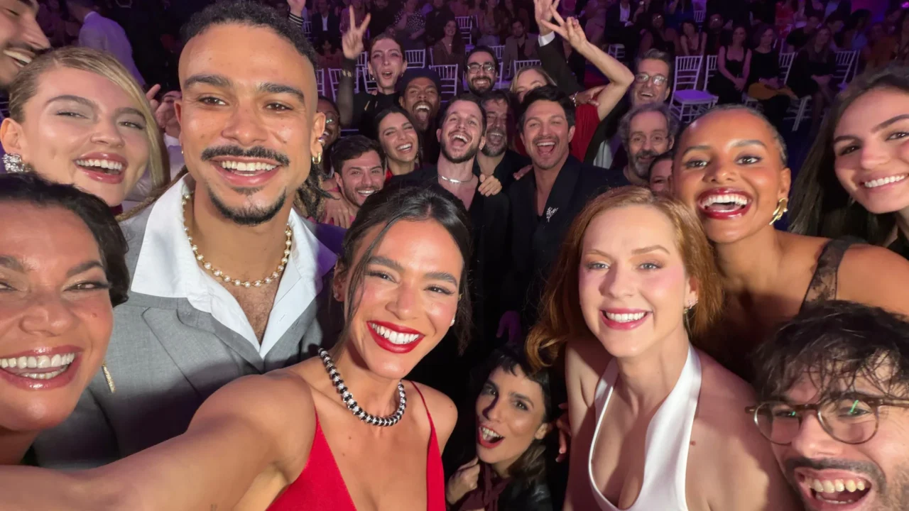 Grupo de pessoas se diverte em evento de première, com decoração em tons de roxo e iluminação de festa, usando roupas elegantes em cores como vermelho, branco e preto