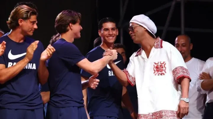 Homem de chapéu branco e camisa rendada vermelha e branca cumprimenta jovem de camiseta azul-marinho com logo Nike, enquanto outros aplaudem em palco