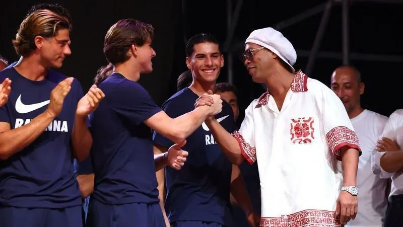 Homem de chapéu branco e camisa rendada vermelha e branca cumprimenta jovem de camiseta azul-marinho com logo Nike, enquanto outros aplaudem em palco