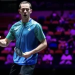 Atleta de camisa azul e verde faz gesto de comemoração com o punho cerrado e uma raquete na mão em arena com arquibancadas ao fundo