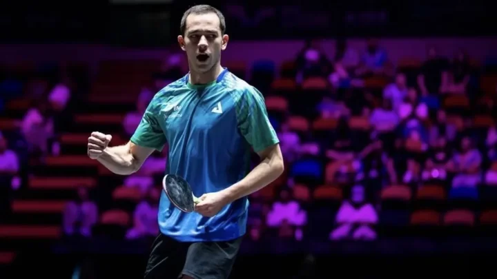 Atleta de camisa azul e verde faz gesto de comemoração com o punho cerrado e uma raquete na mão em arena com arquibancadas ao fundo