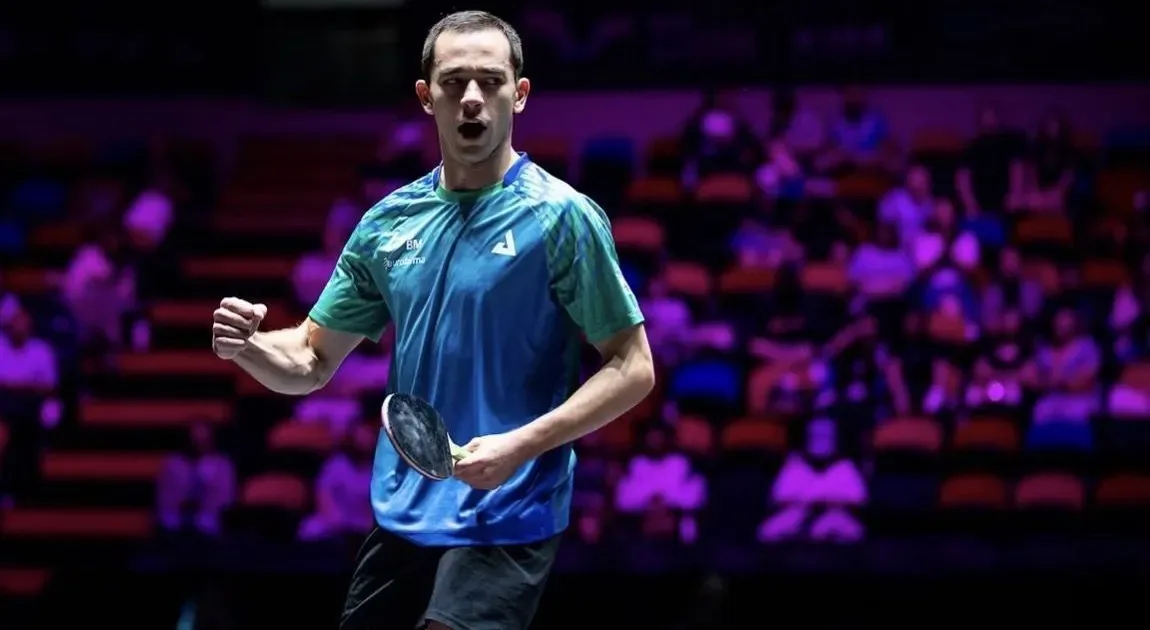 Atleta de camisa azul e verde faz gesto de comemoração com o punho cerrado e uma raquete na mão em arena com arquibancadas ao fundo