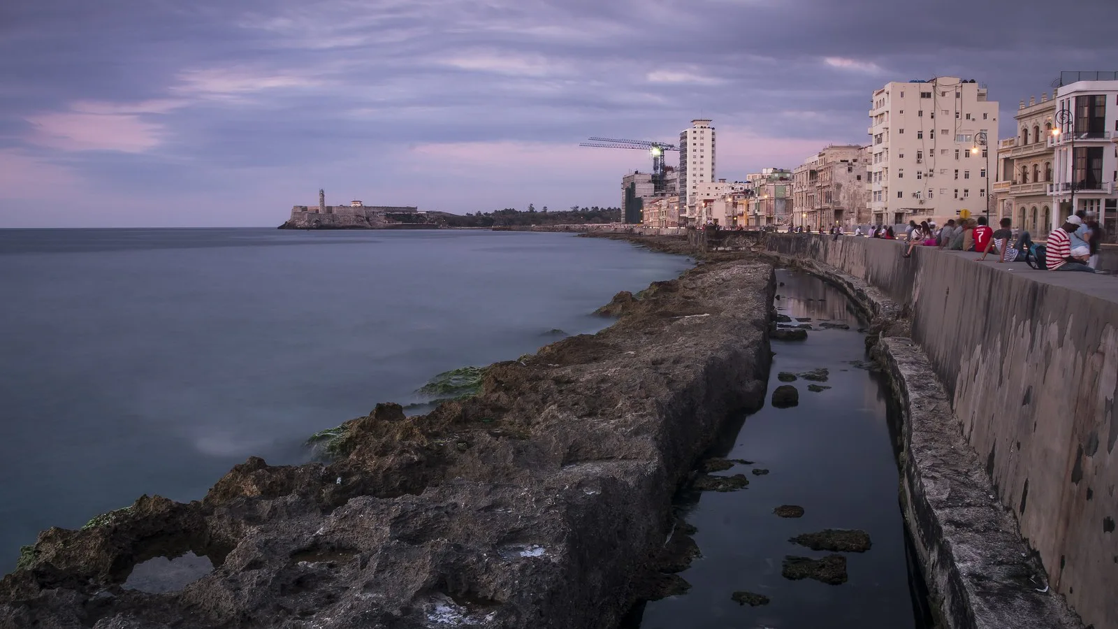 Rochas do litoral junto à mureta do Malecón, em Havana, com prédios à direita e o farol do Castelo do Morro ao fundo, ao entardecer, em abril de 2018 (imagem de arquivo)