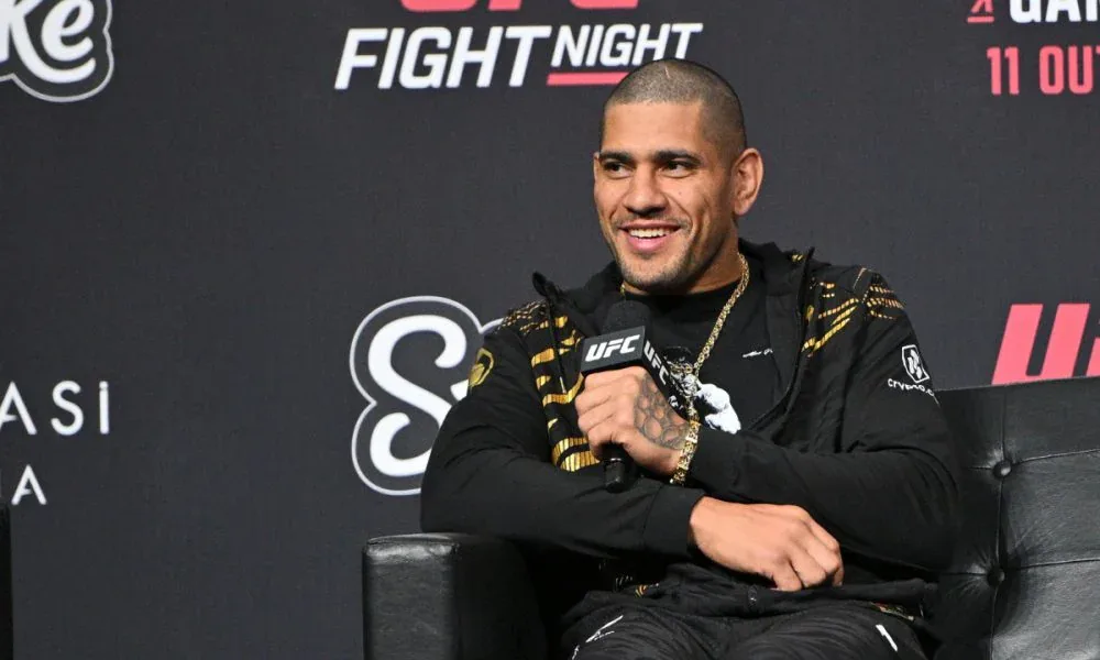 Homem sorridente, de moletom preto com detalhes dourados e corrente no pescoço, segura um microfone do UFC enquanto fala sentado em uma poltrona de couro diante de painel com logotipos do evento