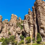 Paredão de torres de pedra de riolito, altas e estriadas, erguidas lado a lado sobre uma encosta com arbustos verdes, sob céu azul, no Chiricahua, no sudeste do Arizona, em 2024